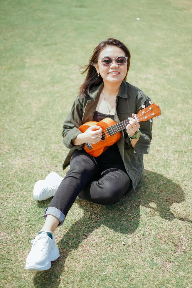 spielen Ukulele von jung schön asiatisch Frau tragen Jacke und schwarz Jeans posieren draußen foto