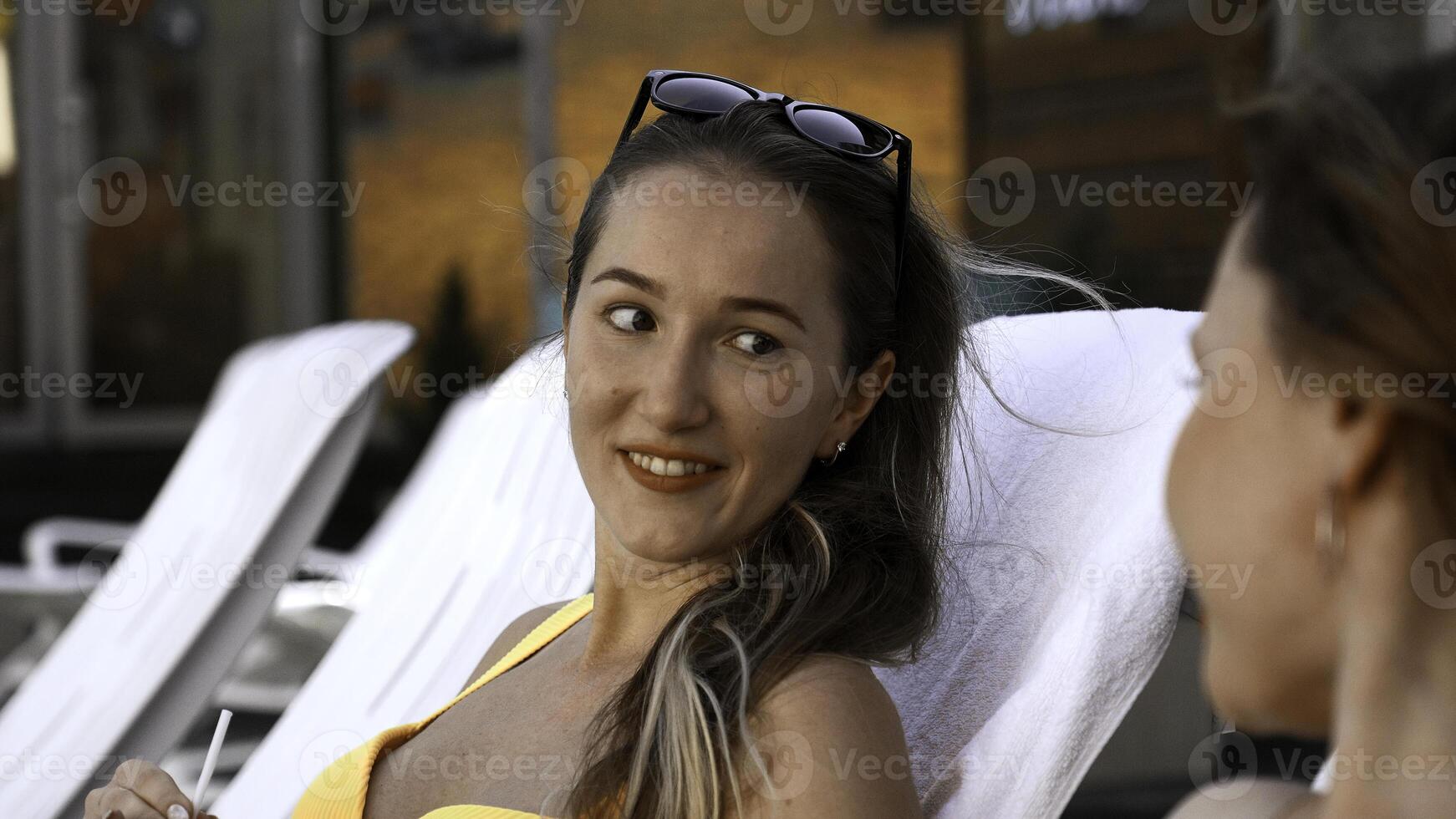glücklich jung Frauen Lügen auf Sonnenliegen durch das Schwimmen Schwimmbad und Klatschen. Clip. entspannend in der Nähe von Resort Schwimmen Schwimmbad mit ein Cocktail. foto