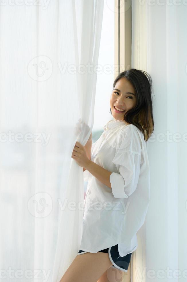 junge asiatische frau im schlafzimmer mit weißen vorhängen und der morgendlichen atmosphäre, wenn die sonne scheint. foto
