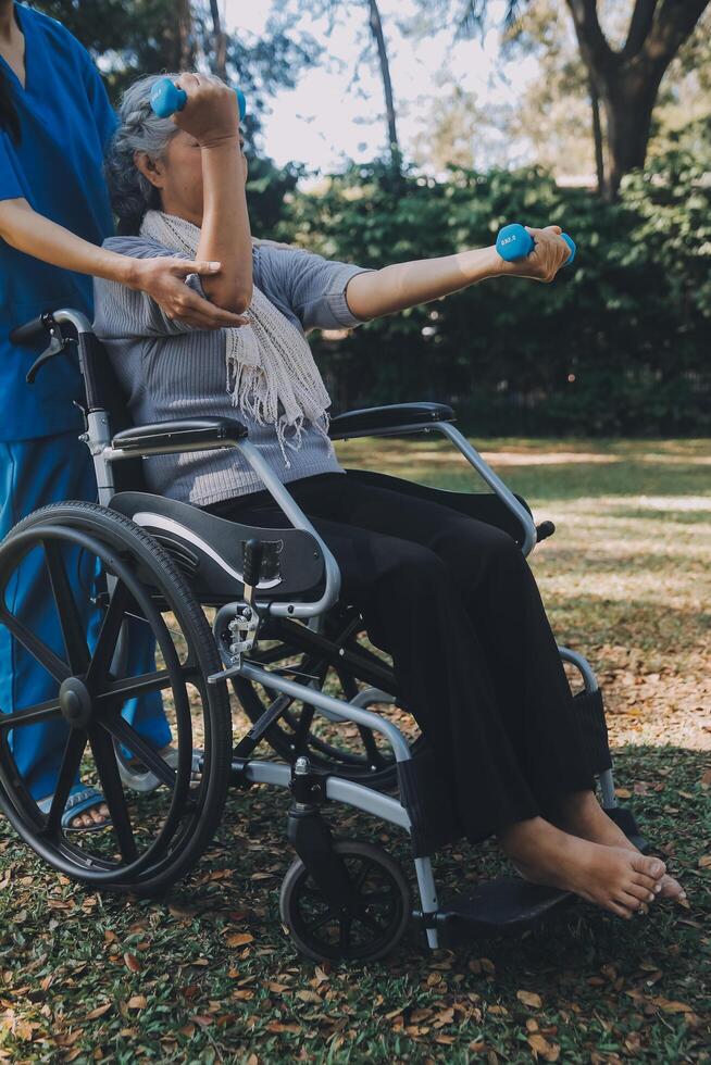 jung Krankenschwester oder Physiotherapeut im Peelings Portion ein glücklich im Ruhestand alt Frau tun Fitness Übungen mit Licht Gewicht Hanteln beim heim. Konzept von Physiotherapie zum Senioren foto