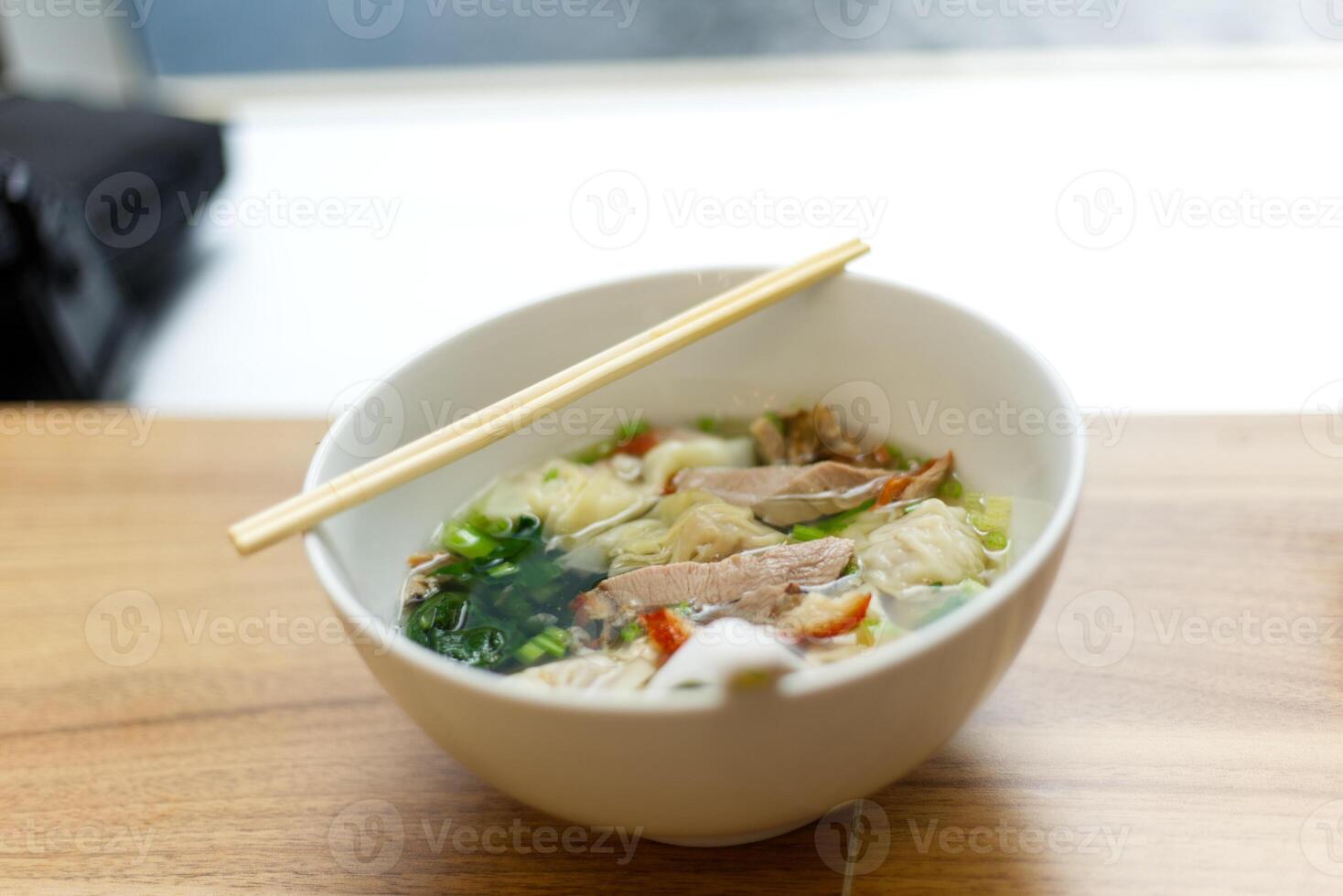 Schweinefleisch Knödel Wonton Suppe mit bok Choy und gegrillt rot Schweinefleisch Dienen mit Essstäbchen auf ein Schüssel auf hölzern Tabelle und Sonnenlicht Hintergrund foto