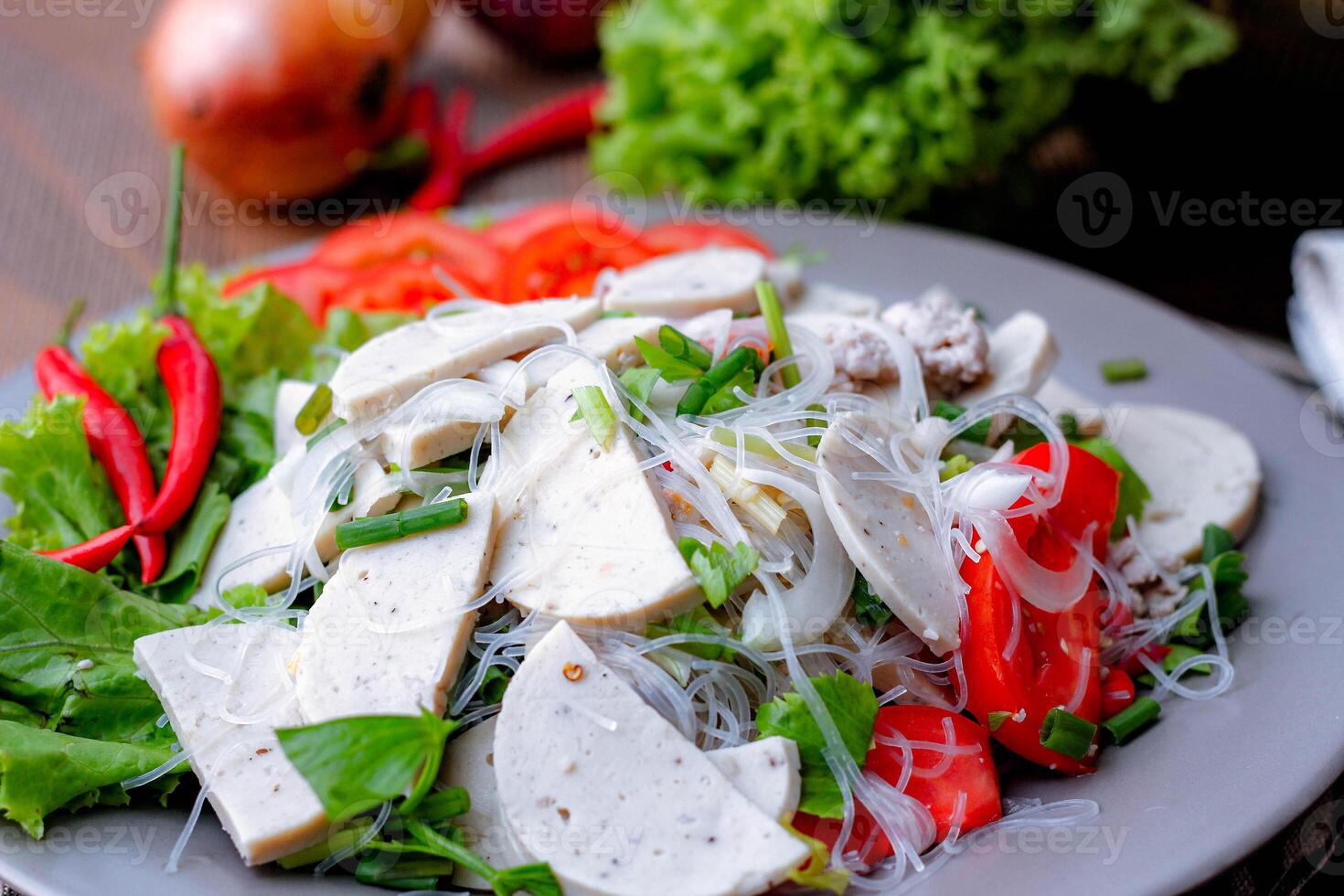 würzig Glas Nudel Salat mit Vietnamesisch Würstchen serviert im ein grau Teller stellen auf ein hölzern Tabelle und verschiedene Gemüse. foto