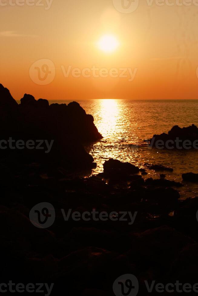 das Sonne ist Rahmen auf das Meer mit Felsen im das Vordergrund, schwarz Silhouetten auf das Felsen, Licht reflektieren aus das Meer. foto