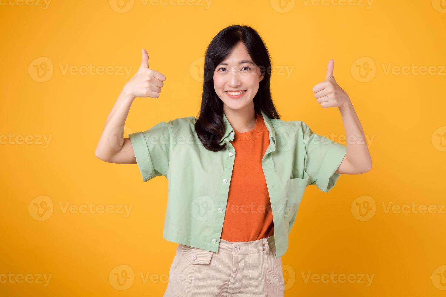 feiern die Genehmigung mit jung asiatisch Frau 30er Jahre, tragen im Orange Hemd und Grün Jumper. ihr Daumen oben Geste, isoliert auf beschwingt Gelb Hintergrund, bedeutet ein Konzept von Positivität und Vereinbarung. foto