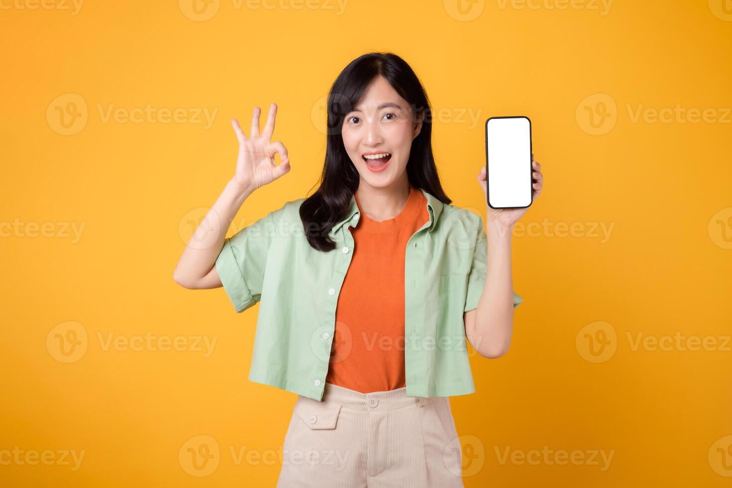 Neu Handy, Mobiltelefon Anwendung mit ein beschwingt jung asiatisch Frau im ihr 30er Jahre, gekleidet im Orange Hemd und Grün Jumper, präsentieren Smartphone Bildschirm mit ein okay Hand Geste auf Gelb Studio Hintergrund. foto