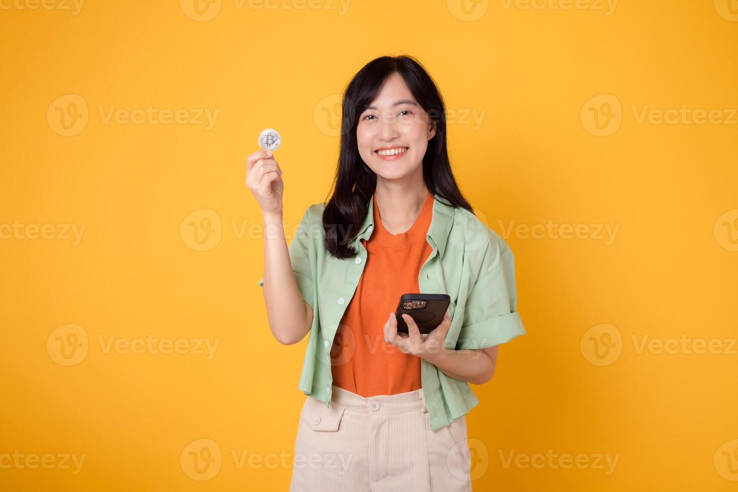 glücklich jung asiatisch Frau im ihr 30er Jahre, tragen Orange Hemd und Grün Jumper, Vitrinen Krypto Währung Münze während halten Smartphone auf Gelb Hintergrund. Zukunft Finanzen Konzept. foto