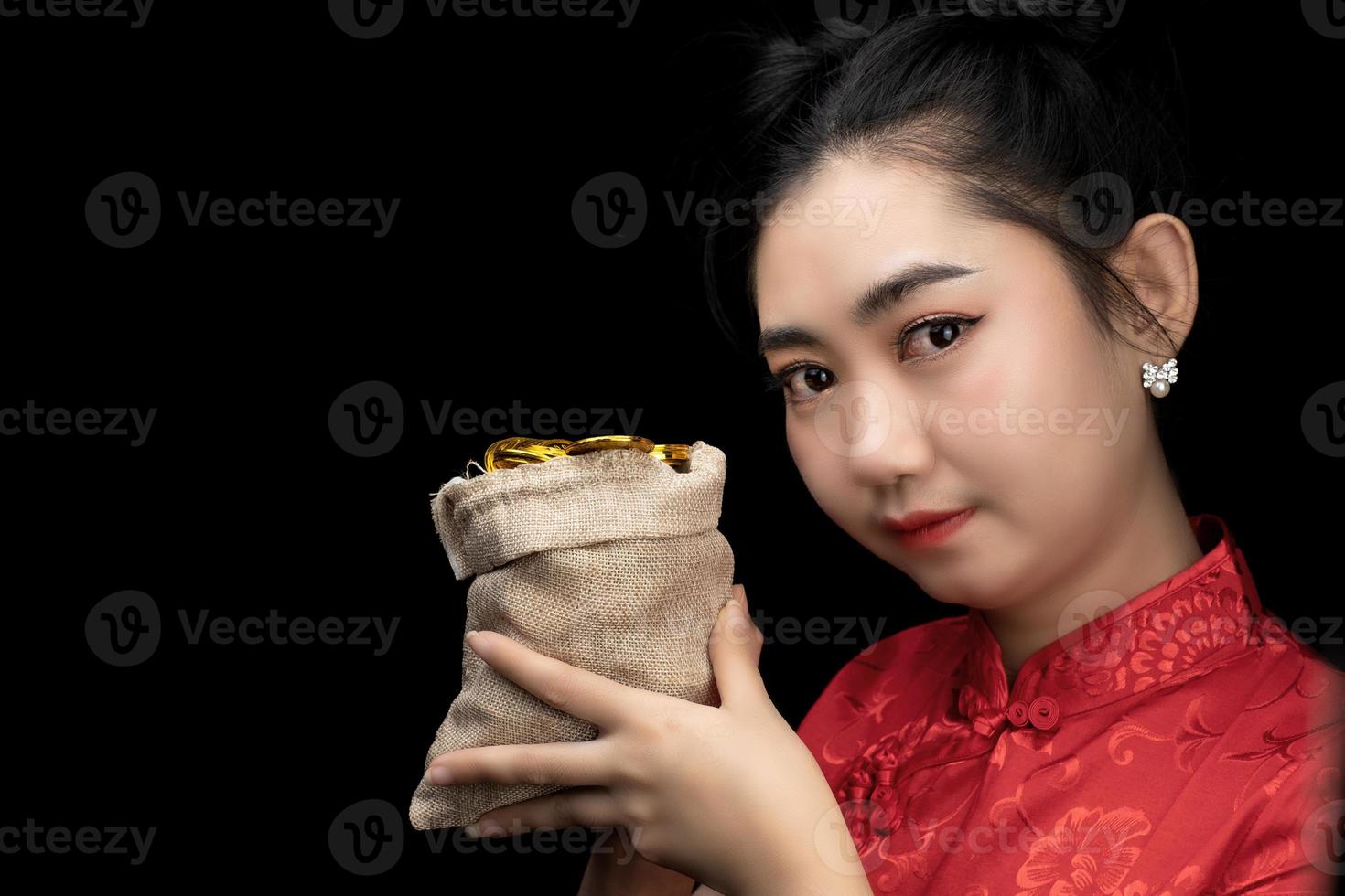 Porträt asiatische junge Frau rotes Kleid traditionelles Cheongsam hält eine Goldmünze in einem Sack auf dem schwarzen Hintergrund foto