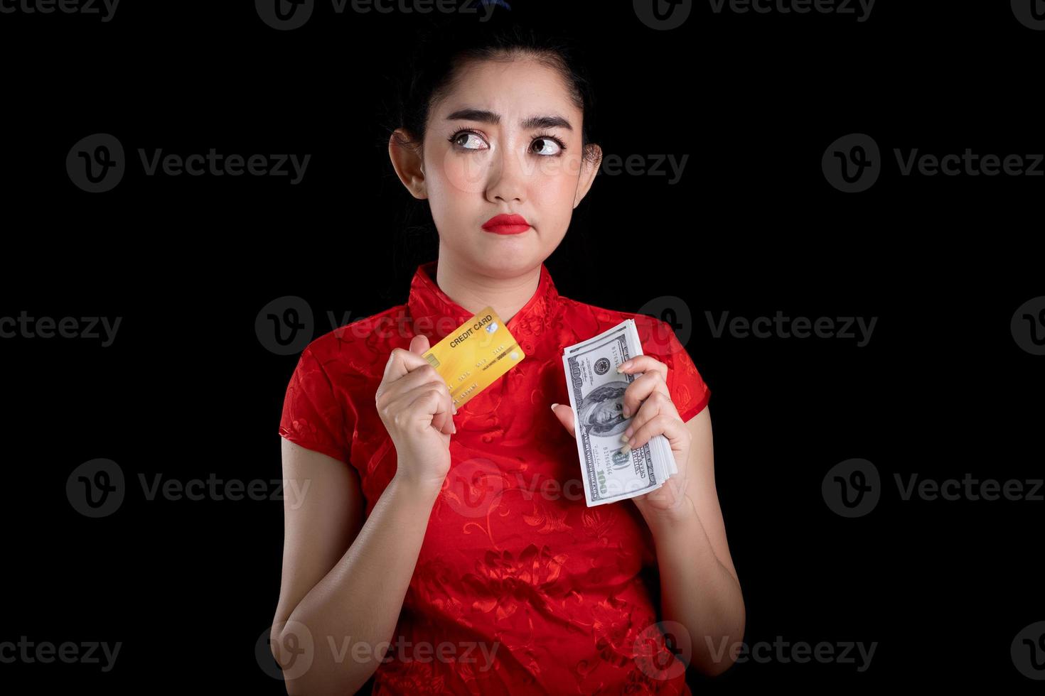 Porträt einer schönen jungen asiatischen Frau, rotes Kleid, traditionelles Cheongsam, das Kreditkarte und Geldbanknote 100 usd am schwarzen Hintergrund hält foto