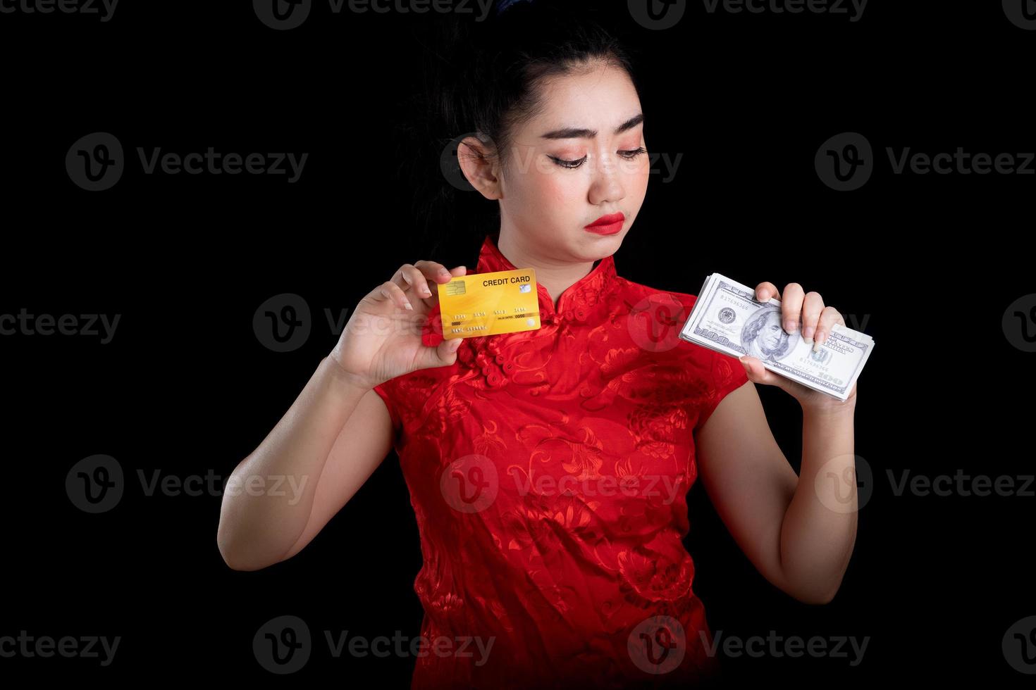 Porträt einer schönen jungen asiatischen Frau, rotes Kleid, traditionelles Cheongsam, das Kreditkarte und Geldbanknote 100 usd am schwarzen Hintergrund hält foto