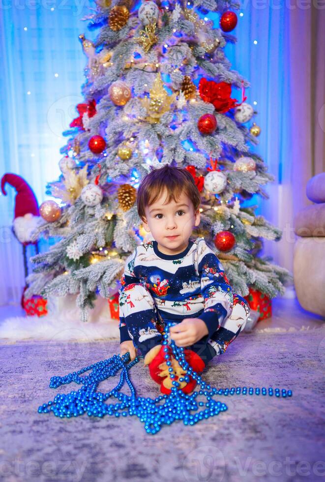 ein froh wenig Junge bewundern das festlich Weihnachten Baum. ein wenig Junge Sitzung im Vorderseite von ein Weihnachten Baum foto