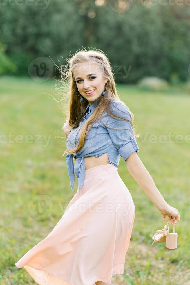 jung kaukasisch Frau oder Mädchen im ein Sommer- Kleid und ein Denim Hemd ist Gehen auf das Gras, halten Schuhe im ihr Hände, entspannend draußen das Stadt auf ein Wochenende im Sommer- foto