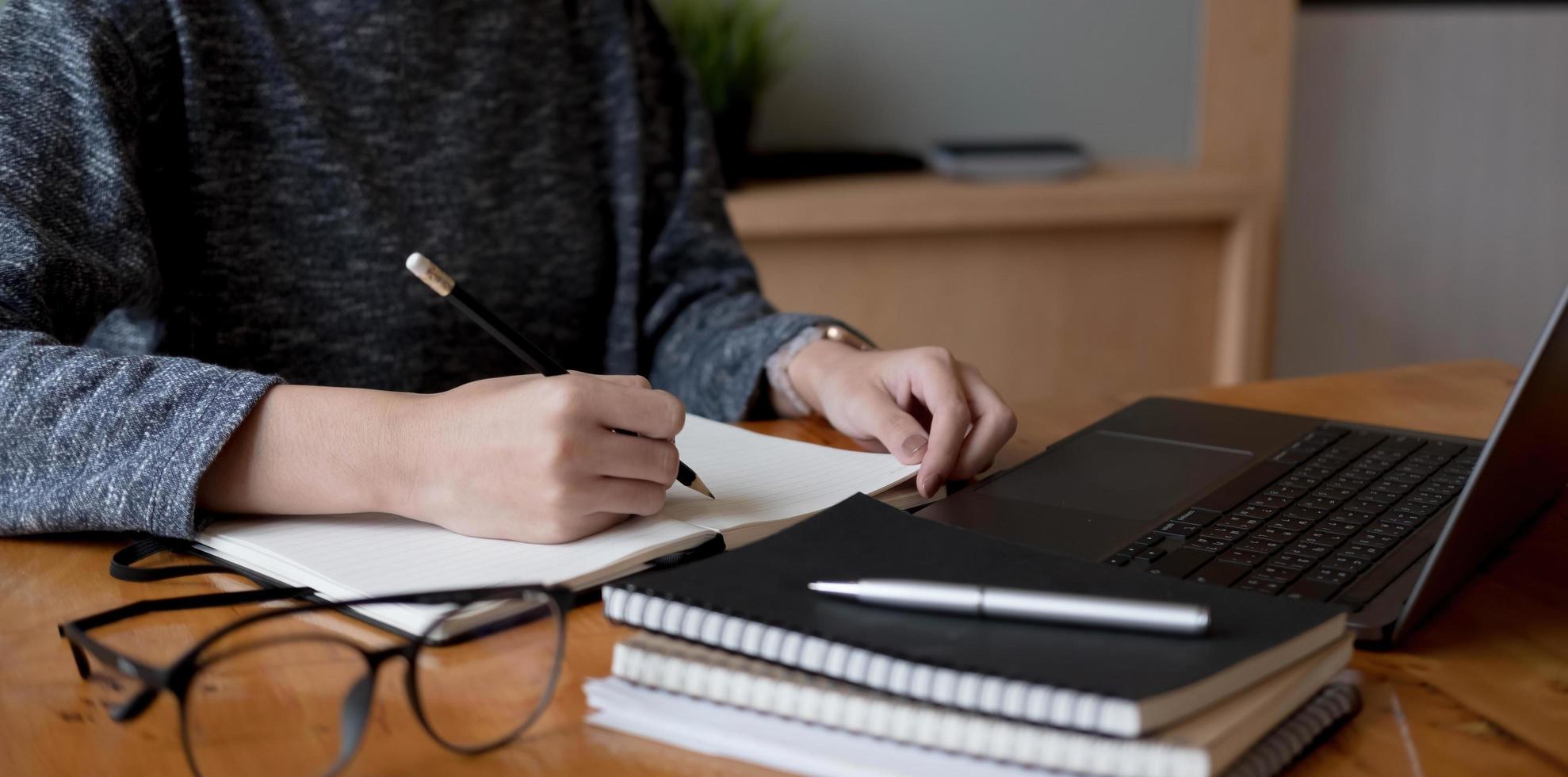 abgeschnittene Fotohand einer Frau, die eine Liste schreibt und Notizen im Notizblock macht, die zu Hause mit dem Laptop online arbeitet oder lernt foto