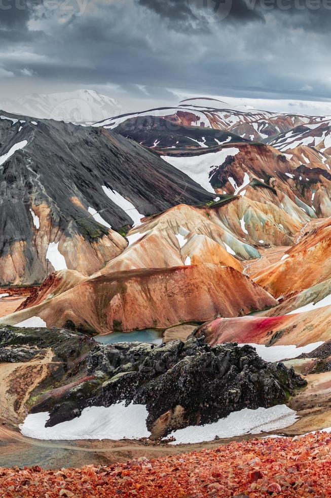 erstaunliche isländische Landschaft mit farbenfrohen regenbogenfarbenen vulkanischen Landmannalaugar-Bergen, am berühmten Laugavegur-Wanderweg mit dramatischem Schneehimmel und rotem Vulkanboden in Island foto