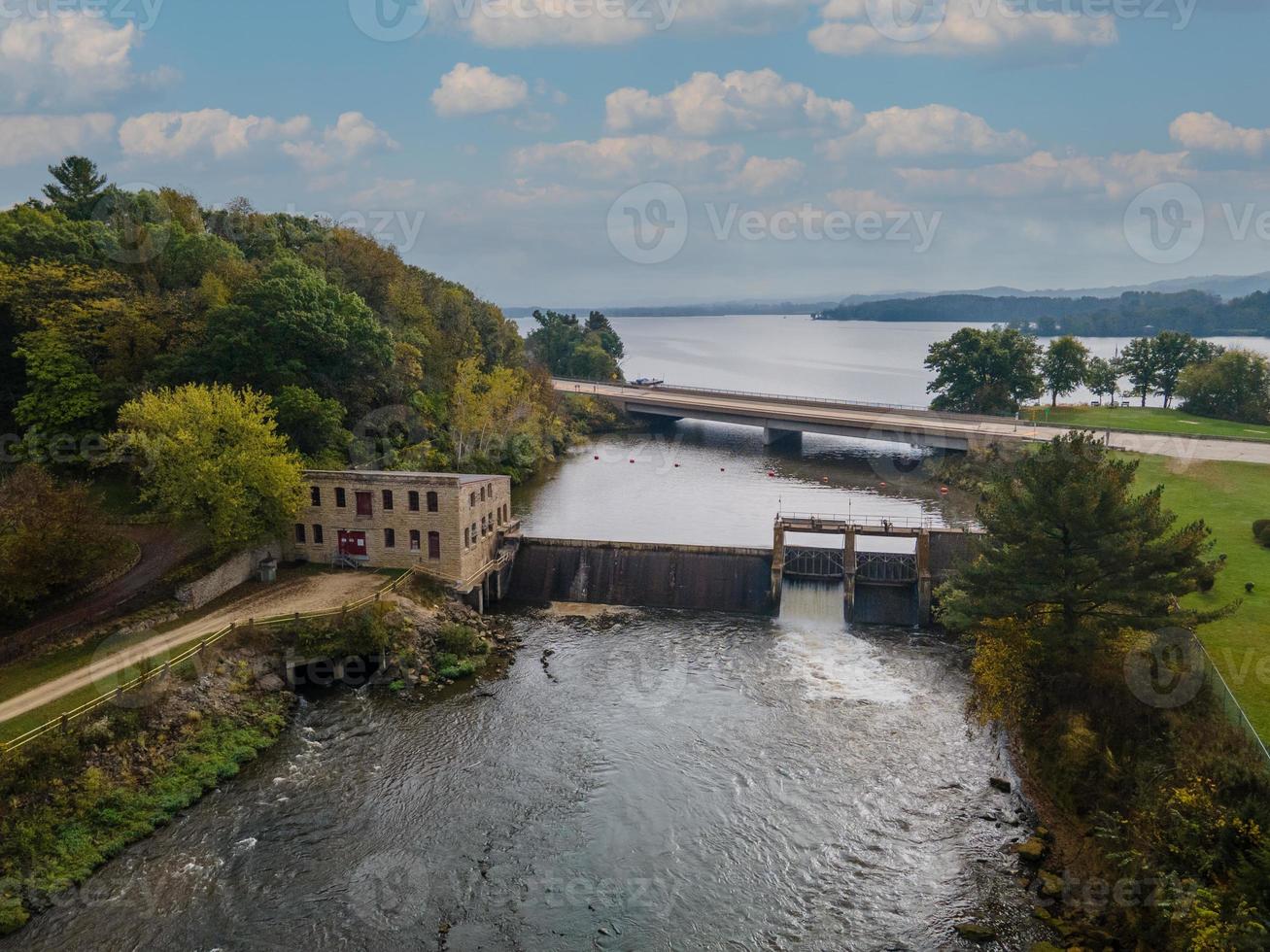 Luftaufnahme des Damms mit Brücke über den Fluss zum See im Herbst, historisches Steuerhaus mit Schotterstraße, aktiver Überlauf foto