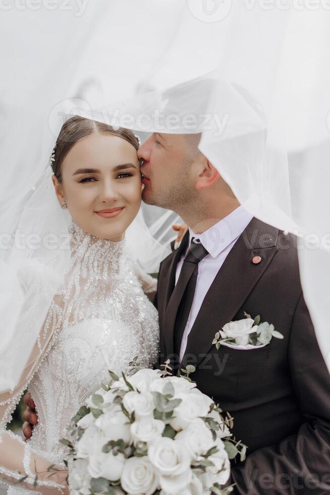 das Braut im ein elegant Kleid mit ein Strauß und das Bräutigam im ein klassisch passen sind Gehen im das Garten, halten Hände. foto