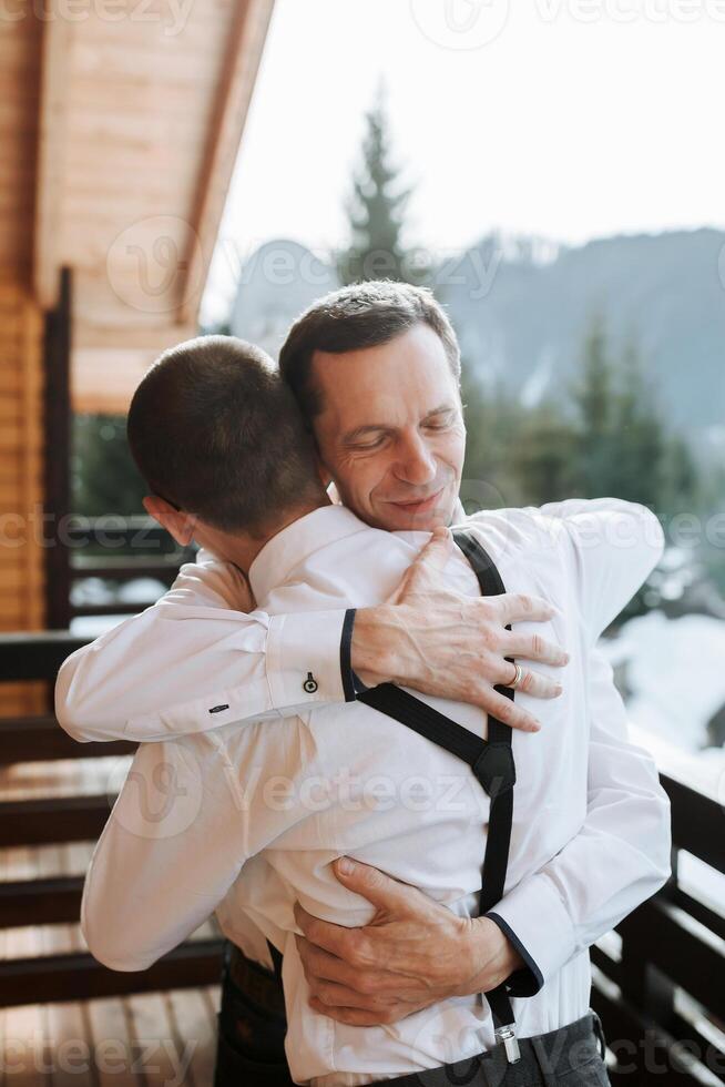 ein Vater Umarmungen seine reifen Sohn und hilft ihm bereiten zum das Hochzeit Zeremonie. warm und aufrichtig Beziehungen zwischen ein Vater und ein Erwachsene Sohn. ein emotional Moment beim ein Hochzeit foto