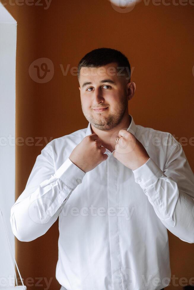 das Bräutigam Tasten seine Hemd durch das Fenster. Mann im ein Shirt. männlich Porträt. Fotoshooting beim das Fenster. Bräutigam Sammlung. Hochzeit Tag. gut aussehend Mann Bräutigam Knöpfen seine Hemd im ein Hotel Zimmer. foto