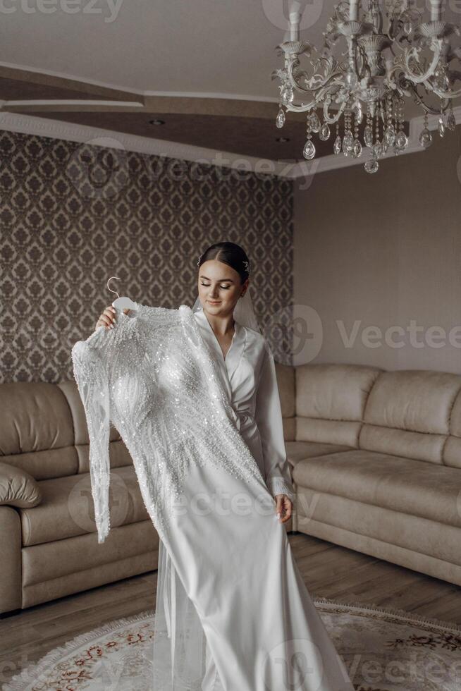 das Braut ist halten ihr exquisit Hochzeit Kleid und posieren im ihr Zimmer. Morgen von das Braut. Porträt foto