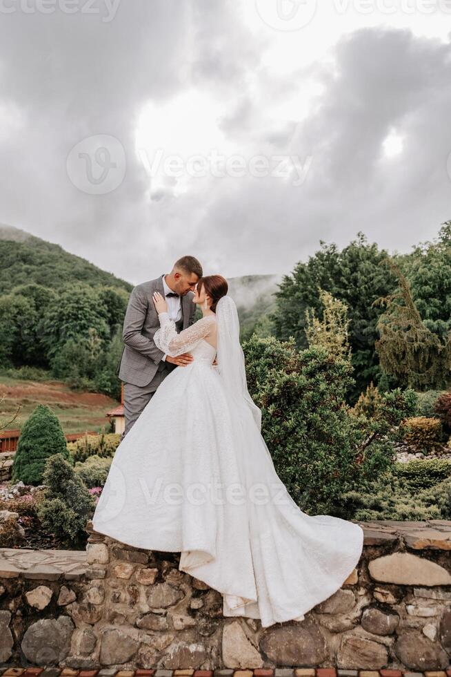 Hochzeit. Liebe und Paar im Garten zum Hochzeit. Feier von Zeremonie und Engagement. speichern das Datum. Vertrauen. das Bräutigam umarmt das Braut gegen das Hintergrund von Berge und ein wolkig Himmel. foto