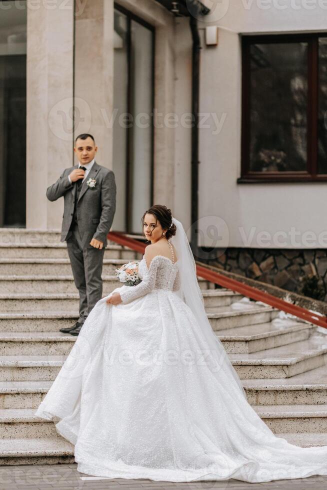 ein Braut im ein Weiß Kleid mit ein Zug und ein Bräutigam im ein passen Pose auf das Schritte von ein Gebäude. Hochzeit Foto Session im Natur