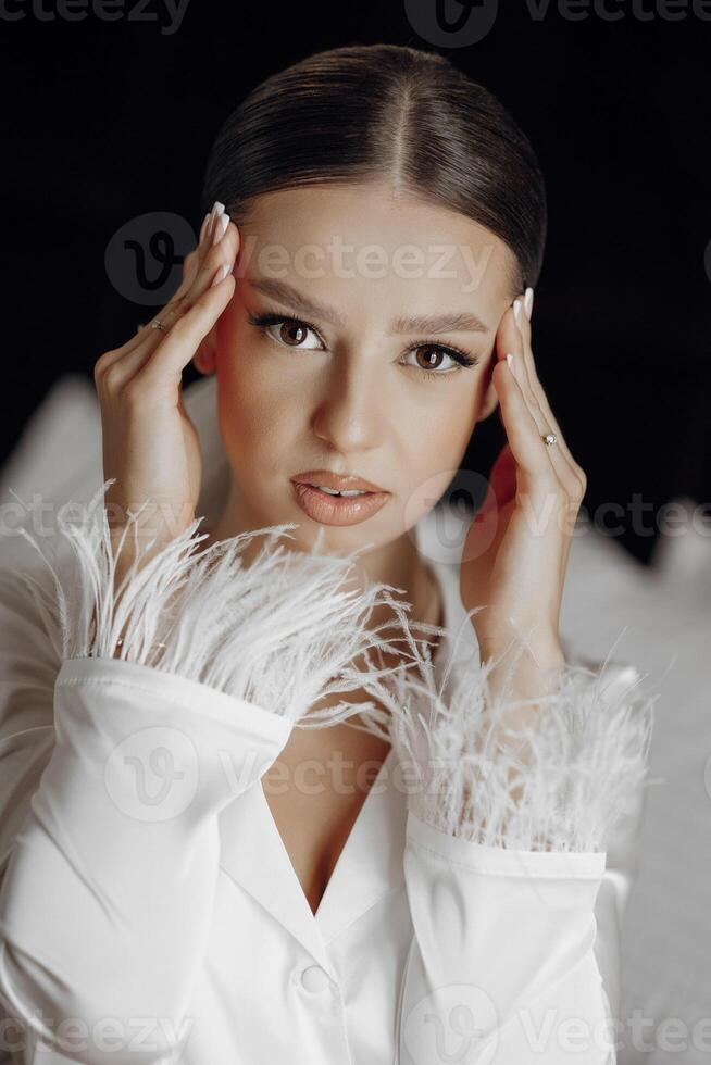 Porträt von glamourös und zuversichtlich Braut auf Bett im Weiß Pyjama im Hotel Zimmer. modern Haar und bilden. Hochzeit Konzept. Morgen von das Braut. Vorbereitung zum das Hochzeit Zeremonie foto