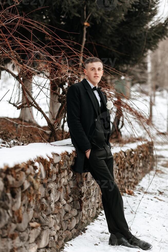 Hochzeit Porträt von das Bräutigam. das Bräutigam ist Stehen in der Nähe von ein Stein Zaun. ein Mann im ein schwarz Anzug. Winter Hochzeit foto