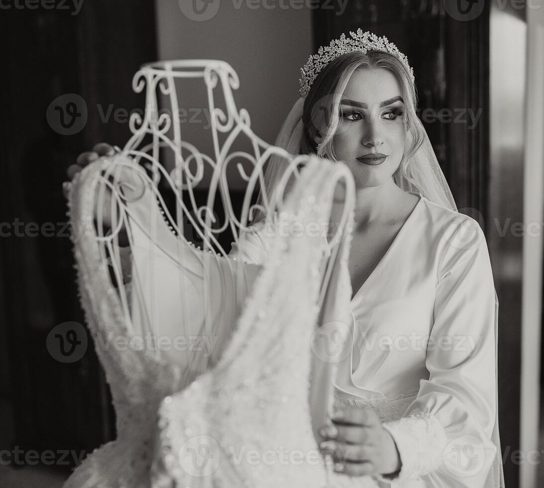 ein Braut im ein Seide passen mit ein Schleier posiert im ihr Zimmer Nächster zu ihr Hochzeit Kleid. Morgen von das Braut. Vorbereitung. schwarz und Weiß Foto