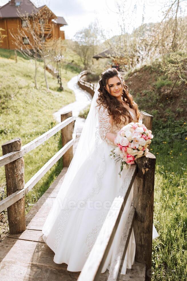 lockig Brünette Braut posiert mit ein Strauß, in der Nähe von ein hölzern Geländer. großartig Kleid mit lange Ärmel, öffnen Büste. Frühling Hochzeit foto