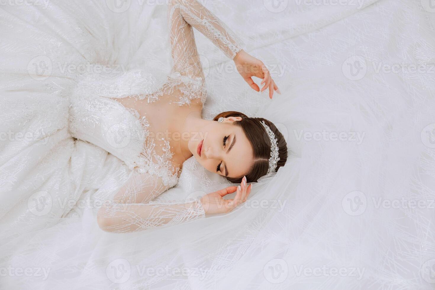 ein Brünette Braut mit ein Tiara im ein Spitzen- Weiß Kleid mit lange Ärmel posiert gegen das Hintergrund von ihr Kleid. auf ein Weiß Hintergrund. klassisch Frisur. Abend bilden. Frühling Hochzeit foto