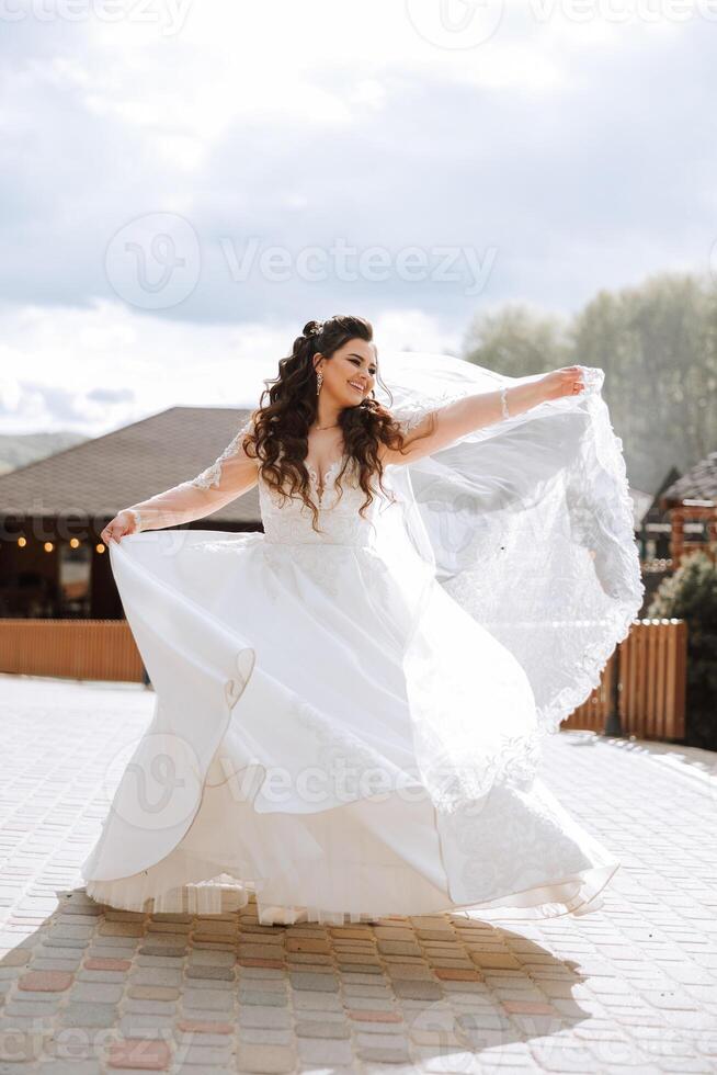 ein lockig Braut wirbelt ihr lange Schleier gegen ein Hintergrund von Berge und hölzern Häuser. großartig Kleid mit lange Ärmel, öffnen Büste. Sommer- Hochzeit foto