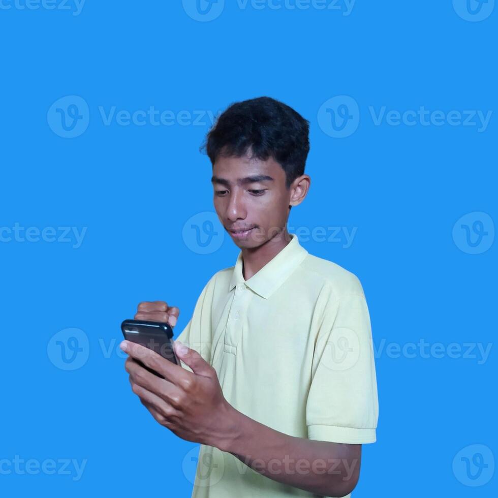 jung asiatisch Mann überrascht suchen beim Clever Telefon tragen Gelb T-Shirt, Blau Hintergrund. foto