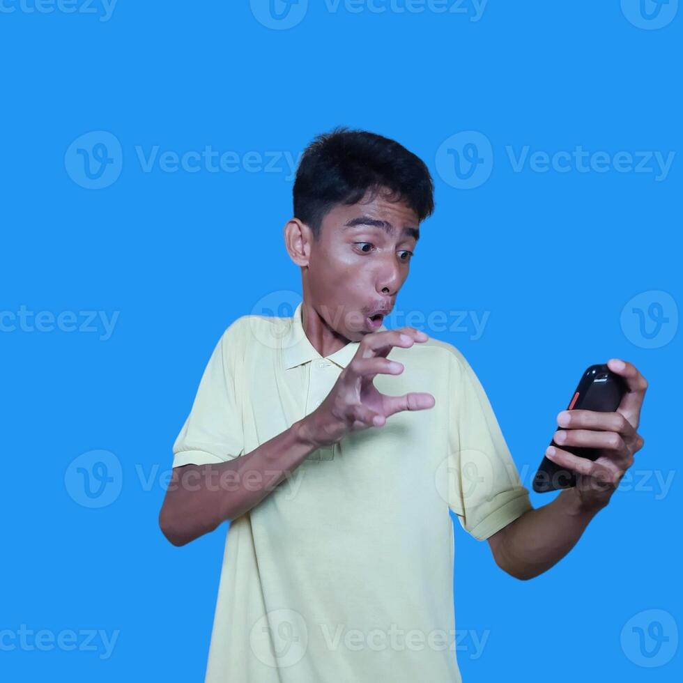jung asiatisch Mann überrascht suchen beim Clever Telefon tragen Gelb T-Shirt, Blau Hintergrund. foto