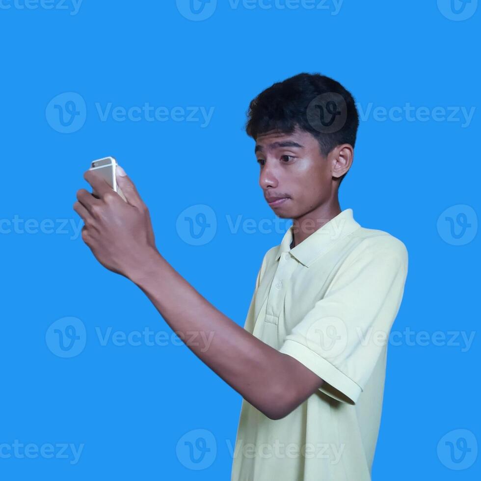 jung asiatisch Mann überrascht suchen beim Clever Telefon isoliert Blau Hintergrund. tragen ein Gelb T-Shirt foto