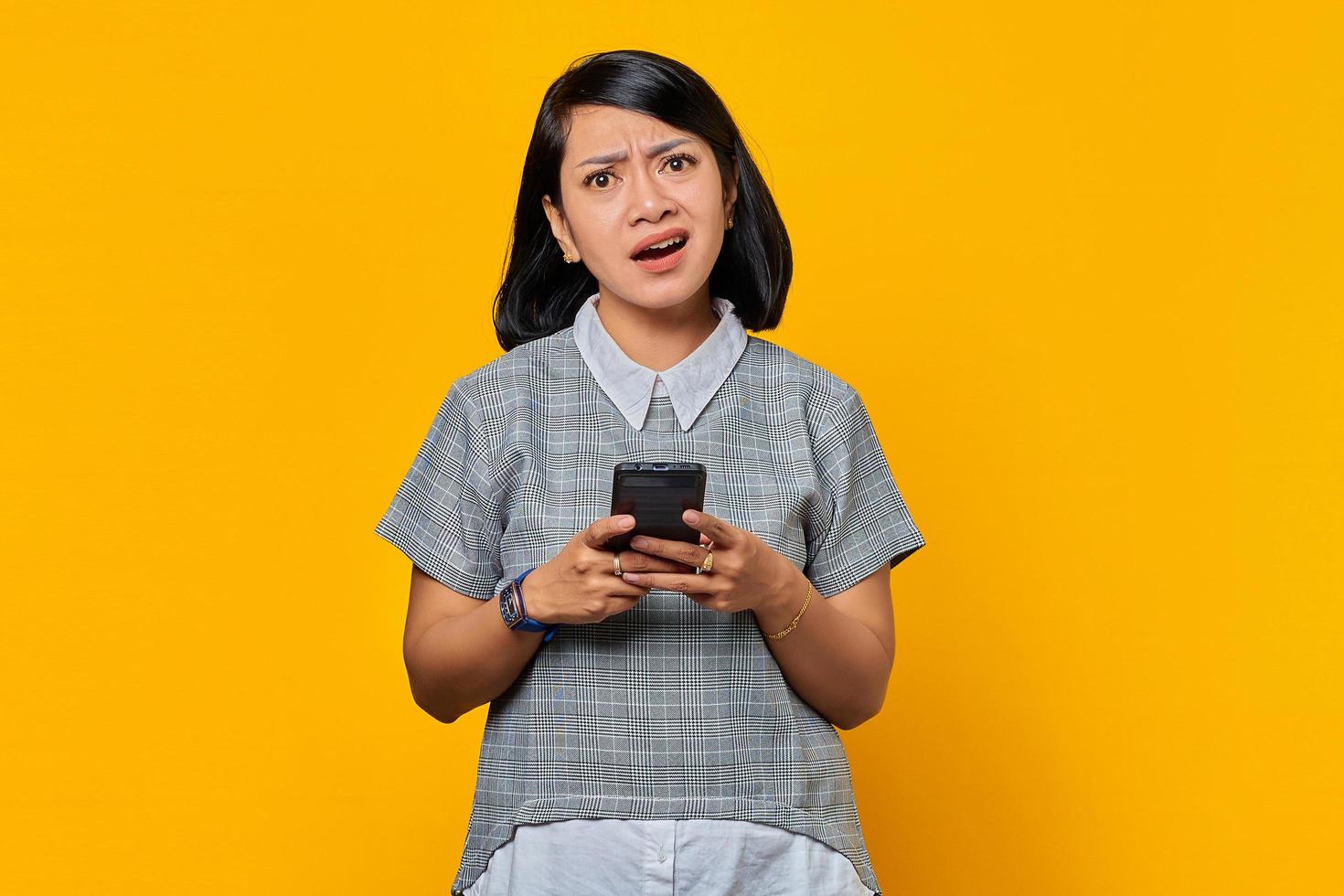 Porträt einer unglücklichen jungen asiatischen frau, die handy hält und die kamera einzeln auf gelbem hintergrund anschaut foto