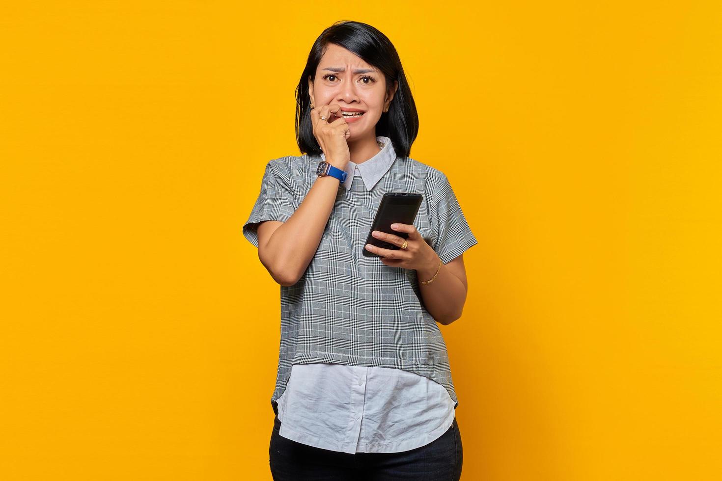 Junge asiatische Frau, die Smartphone hält, sieht ängstlich aus und hat Angst vor etwas, das auf gelbem Hintergrund isoliert ist foto