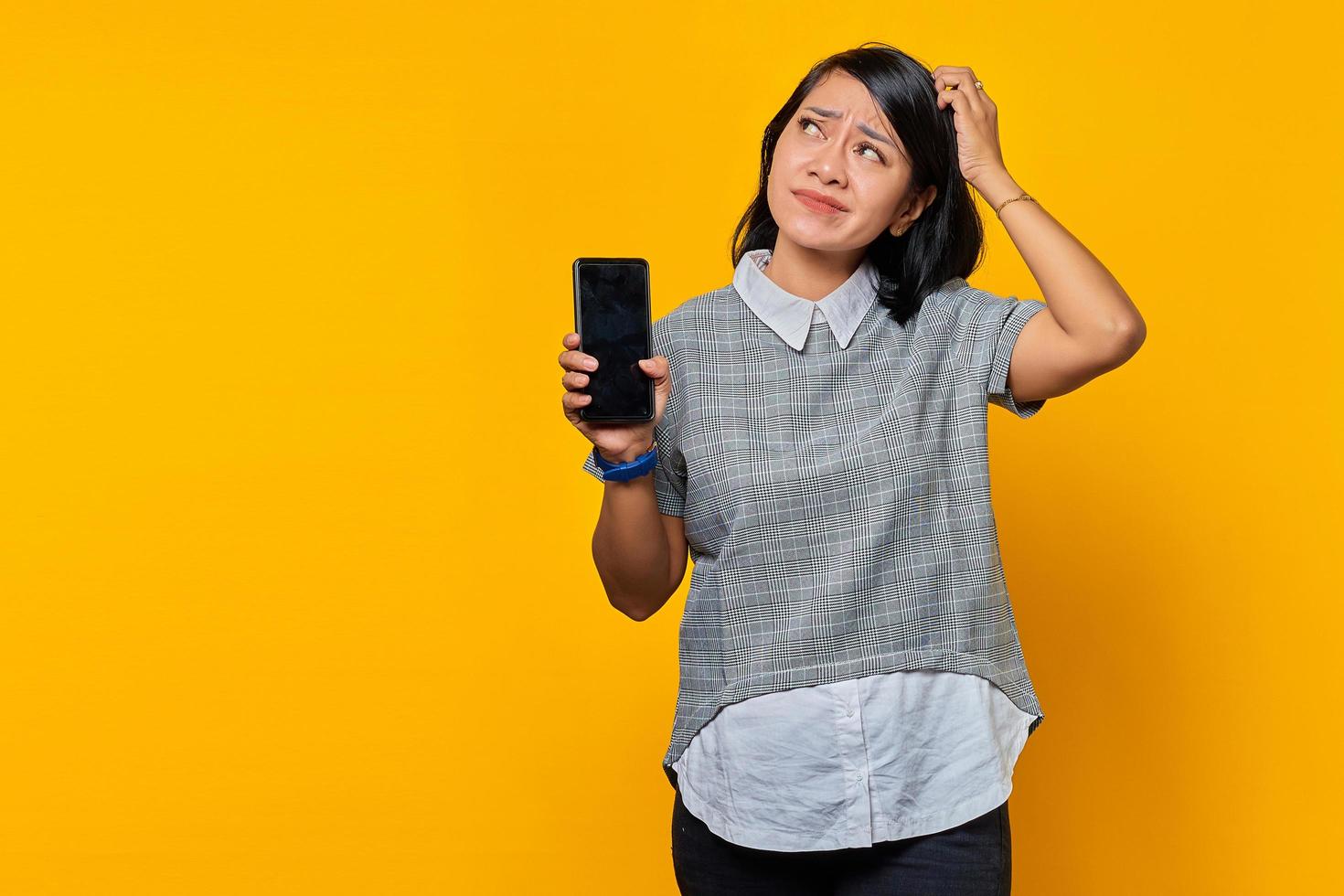 Porträt einer schönen asiatischen Frau, die ein Smartphone hält, ihren Kopf berührt und ihre Besorgnis auf gelbem Hintergrund ausdrückt foto