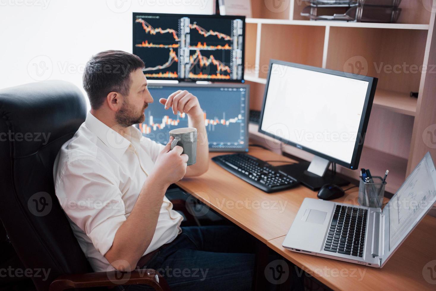 Der junge Geschäftsmann sitzt im Büro am Tisch und arbeitet am Computer mit vielen Monitoren, Diagrammen auf dem Monitor. Börsenmakler analysiert Diagramme für binäre Optionen. Hipster-Mann, der Kaffee trinkt, studiert foto