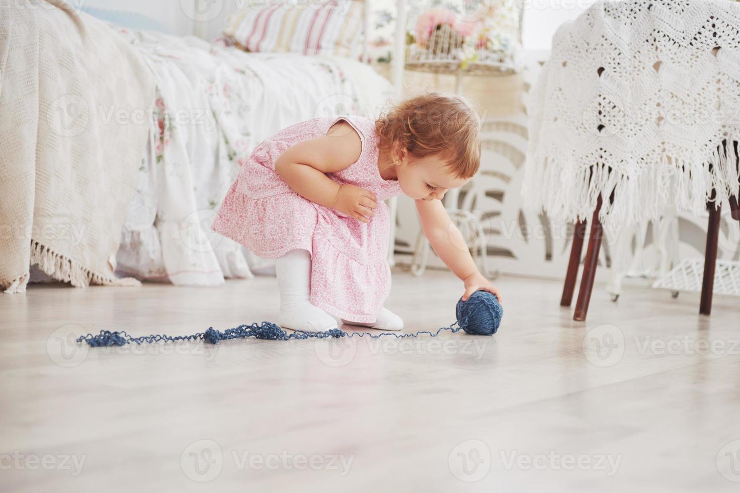 Kindheit Konzept. Babymädchen im niedlichen Kleiderspiel mit farbigem Faden. weißes Vintage-Kinderzimmer foto