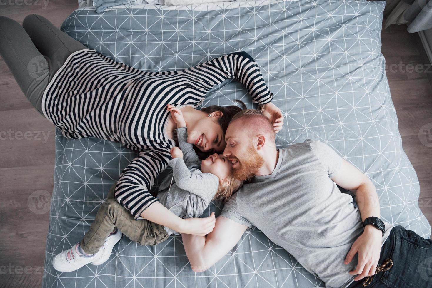 Blick von oben auf die schöne junge Mutter, den Vater und ihre Tochter, die in die Kamera schauen und lächeln, während sie Kopf an Kopf auf dem Bett liegen foto