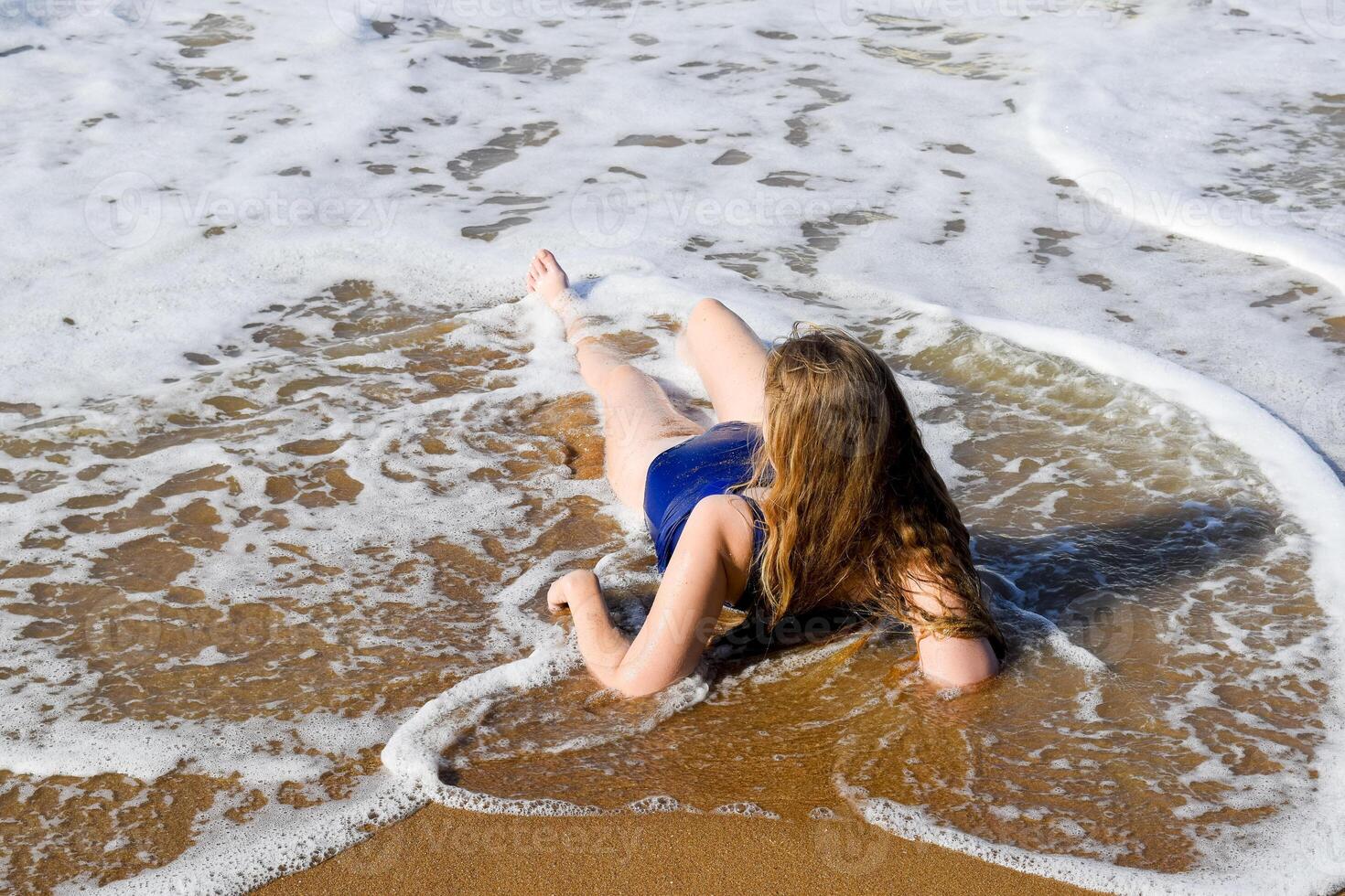 Mädchen im ein Blau Baden passen Lügen auf das Strand und entspannt. Wellen streicheln das Mädchen Körper foto