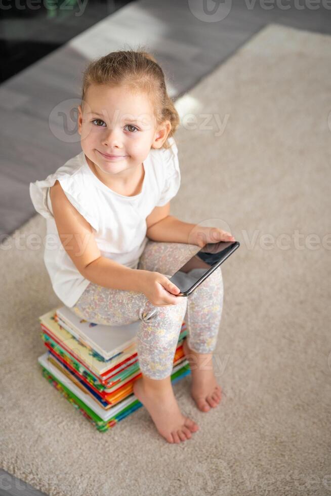 wenig Mädchen sitzt auf Stapel von Kinder- Märchen Bücher mit Smartphone im ihr Hände foto