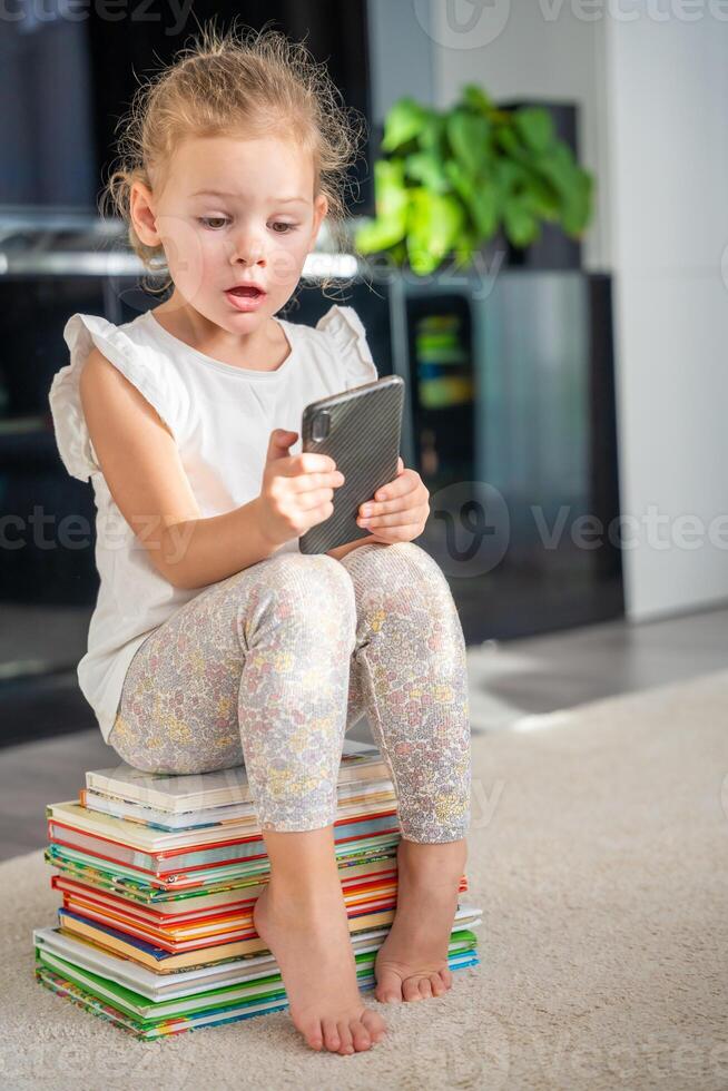 wenig Mädchen sitzt auf ein Stapel von Kinder- Märchen Bücher und Theaterstücke mit Smartphone foto