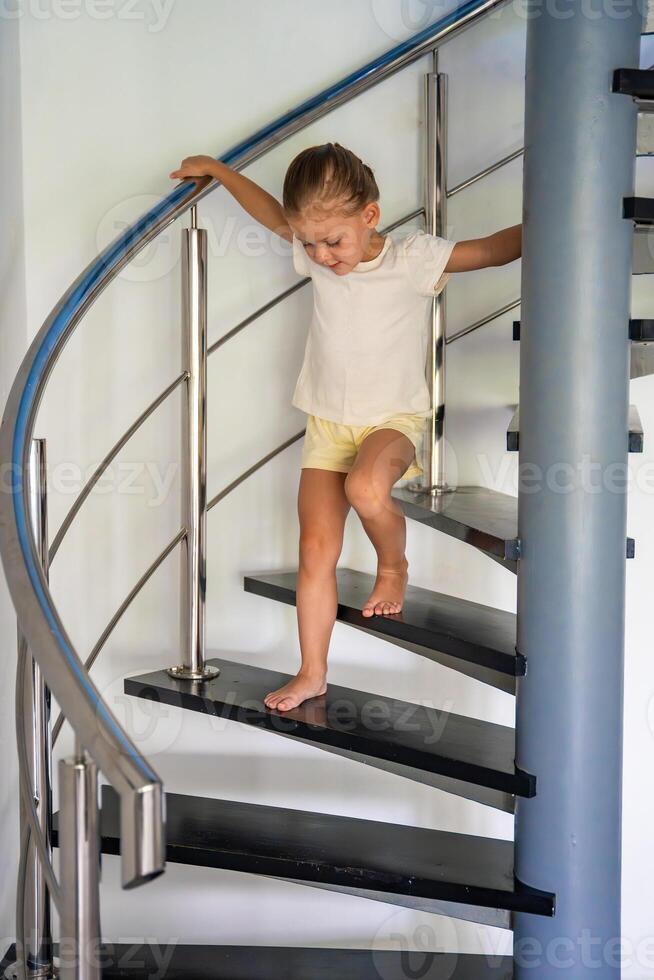 Porträt von wenig Mädchen gehen Nieder das Treppe beim heim, Kind Klettern Spiral- Treppe. gefährlich Situation beim heim. Kind Sicherheit Konzept. foto