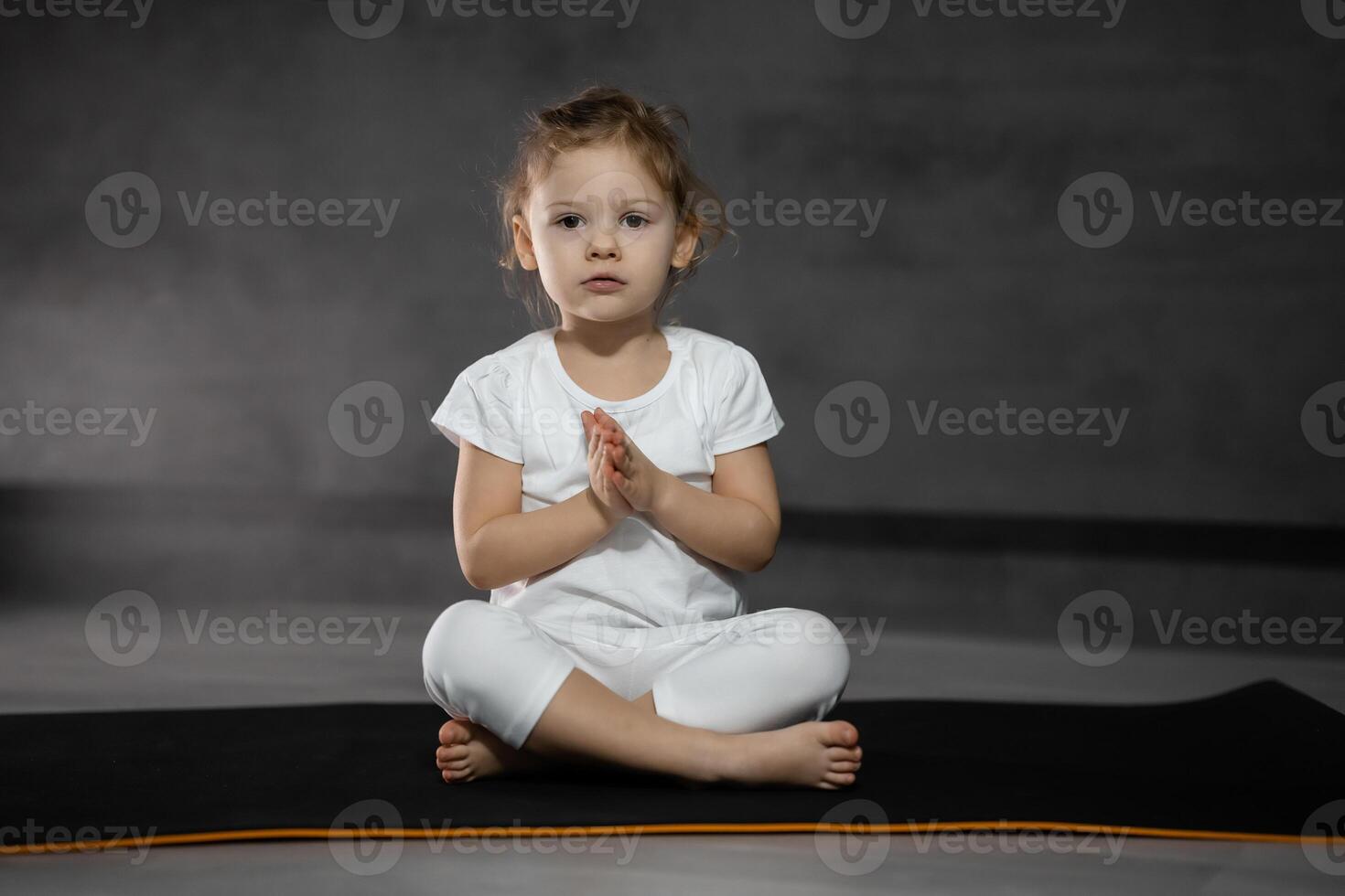 drei Jahre alt wenig Mädchen meditieren im ein Lotus Pose auf ein grau Hintergrund im dunkel Zimmer. hoch Qualität Foto