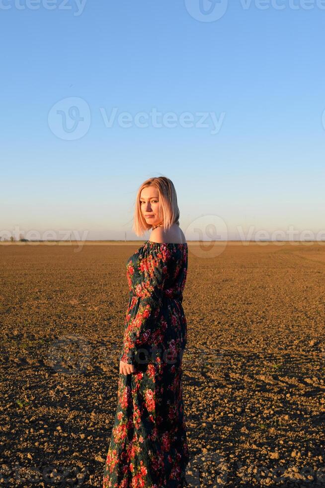 Frau im ein gepflügt Feld im ein rot schwarz Kleid auf ein Sonnenuntergang Hintergrund. foto