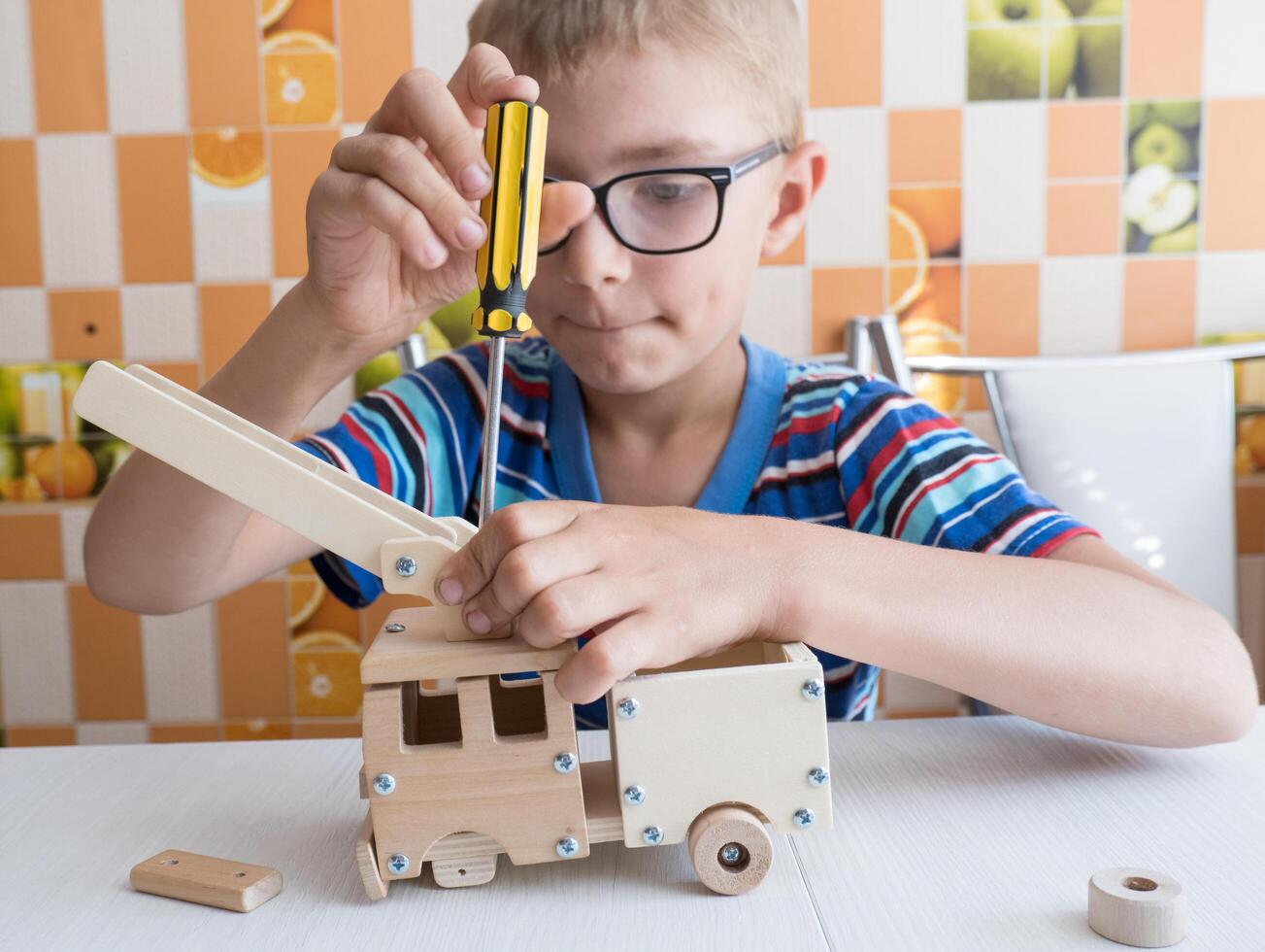 Porträt von ein 7-8 Jahr alt Junge mit ein Schraubendreher, vorsichtig Montage ein hölzern Auto, Sitzung beim das Küche Tisch. Hände schließen oben foto