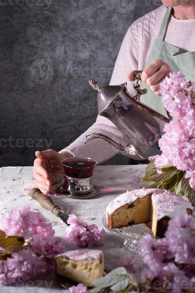 ein Frau gießt Tee von ein Silber Teekanne, Hintergrund von Rosa Sakura Blumen, Ostern foto
