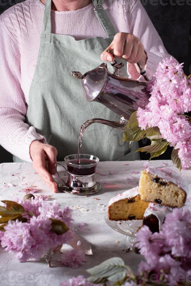 ein Frau gießt Tee von ein Silber Teekanne, Hintergrund von Rosa Sakura Blumen, Ostern foto