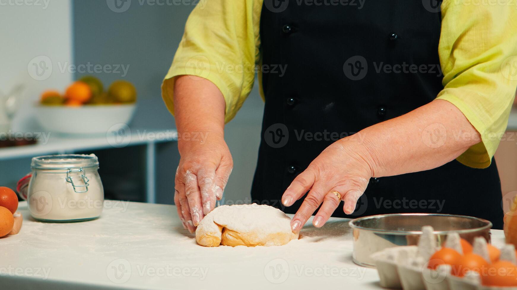 Oma vorbereiten hausgemacht Donuts tragen Küche Schürze. im Ruhestand Senior Koch mit Bonete und Uniform streuen, Sieben Sichtung Weizen Mehl mit Hand Backen hausgemacht Pizza und Brot. foto