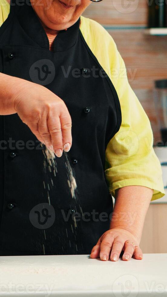 alt Dame vorbereiten Essen im Zuhause Küche Verbreitung Mehl zum Backen ein Rezept. im Ruhestand Alten Koch mit Uniform Prise Sieben Sichtung Zutaten durch Hand auf Tabelle Kochen hausgemacht Pizza Brot foto